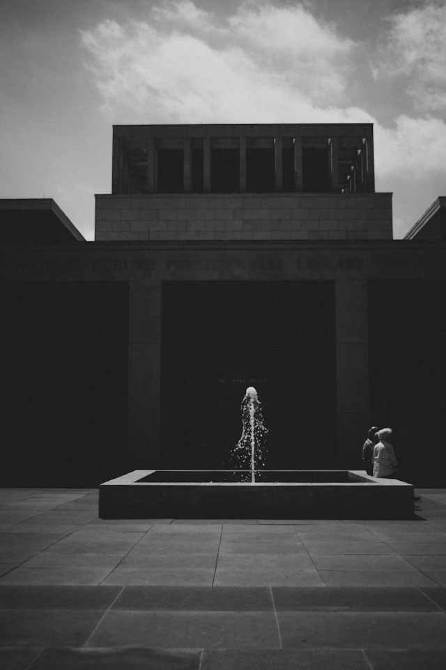 A black and white image of a couple sitting with their back to a fountain as it shoots water straight up.