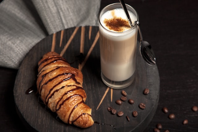 A chocolate bagel and hot drink on a round wooden board with coffee beans on it.